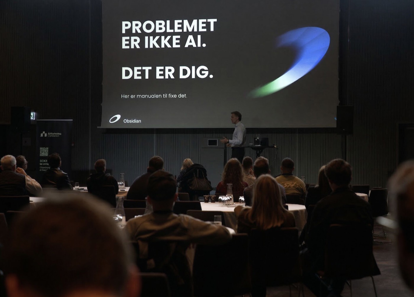 Nicklas Lindby holder AI-foredrag på AI Marketing Conference 2026 foran deltagerne, med sloganet "Problemet er ikke AI. Det er dig" på lærredet